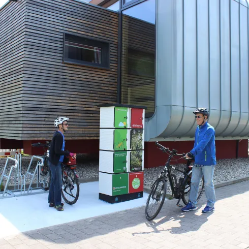 E-Bike Ladestation mit 6 Ladefächern. Davor 2 Radfahrer im Hintergrund das Gebäude des Tourist-Info-Zentrums Pfälzerwald