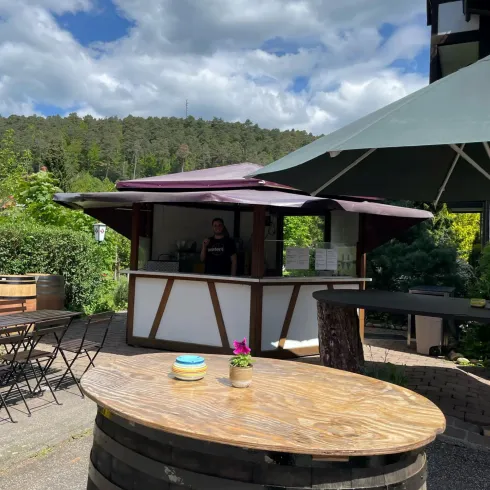 Ein schöner Kiosk mit einer Holzterrasse und einem großen Sonnenschirm. Im Hintergrund sind Bäume und ein blauer Himmel zu sehen.