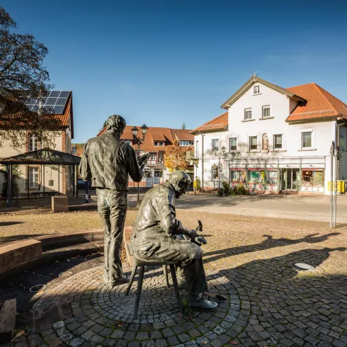 Eine Statue von zwei Männern steht auf einem Platz. Im Hintergrund sind Häuser mit roten Dächern und ein klarer Himmel zu sehen.