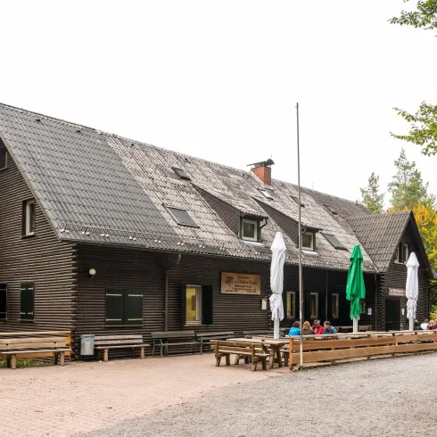 Die Vorderfront des PWV Wanderheims Ortsgruppe Hauenstein e.V. Ein braunes längliches Gebäude eingeschossig. Davor Holztische und- sitzbänke sowie geschlosene Sonnenschirme.