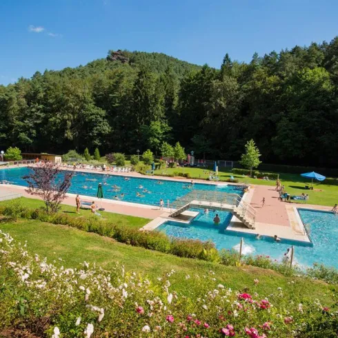 Ein schöner Freibadbereich mit mehreren Schwimmbecken und einer grünen Umgebung. Im Hintergrund sind Bäume und sonnige Himmel zu sehen.