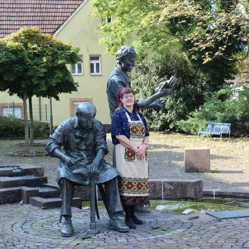 Eine Frau steht zwischen zwei Skulpturen in einem Park. Die Skulpturen zeigen zwei Männer, die anscheinend in einer kreativen oder handwerklichen Tätigkeit beschäftigt sind.