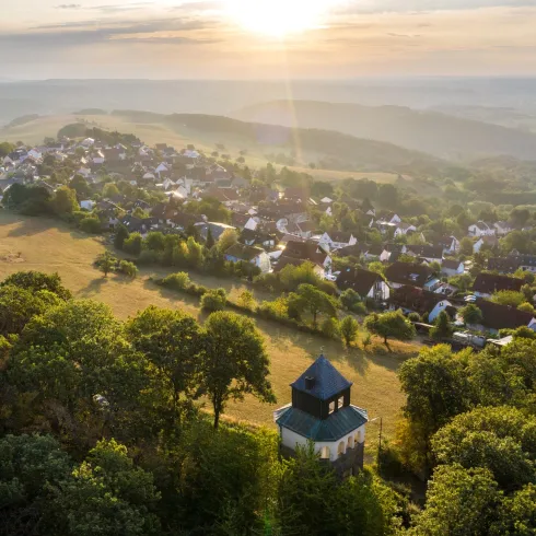 Eine malerische Landschaft mit einem kleinen Dorf im Hintergrund und der Sonne, die über den Horizont aufgeht. Bäume umgeben die Szenerie und schaffen eine ruhige Atmosphäre.