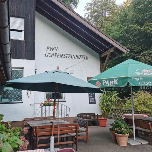 Eine gemütliche Jagdhütte umgeben von grüner Natur. Der Außenbereich ist mit Tischen, Stühlen und einem Sonnenschirm ausgestattet.