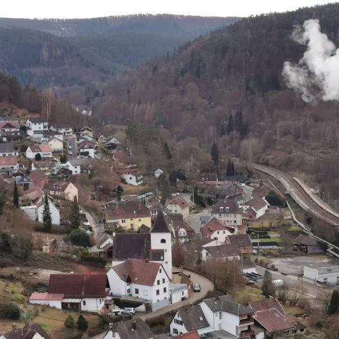 Eine kleine Stadt mit typischen deutschen Häusern und einer rauchenden Industrieanlage. Im Hintergrund sind Hügel und Wälder sichtbar.
