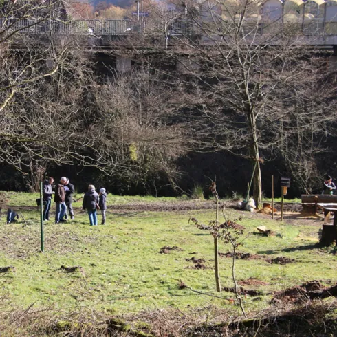 Eine Gruppe von Menschen steht auf einer Wiese umgeben von Bäumen. Im Hintergrund sind Bauwerke und ein kleiner Tischbereich sichtbar.