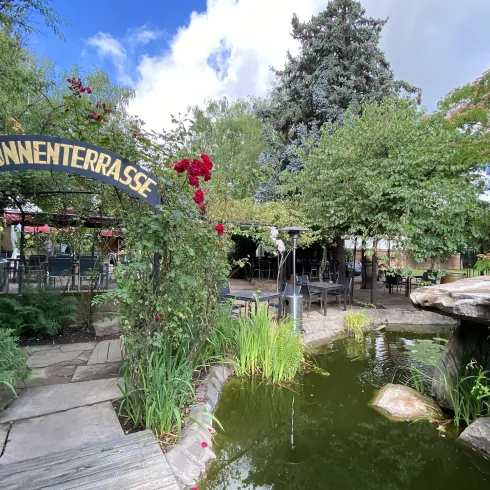 Eine einladende Terrasse mit dem Schriftzug „Brunnenterrasse“ und einem kleinen Teich. Üppiges Grün und Blumen schaffen eine gemütliche Atmosphäre.