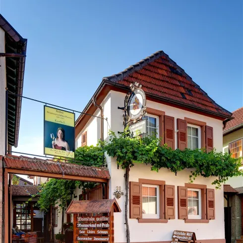 Ein charmantes Gebäude mit einem roten Dach und Fensterläden, umgeben von Weinranken. Die Fassade ist freundlich gestaltet und lädt zum Verweilen ein.