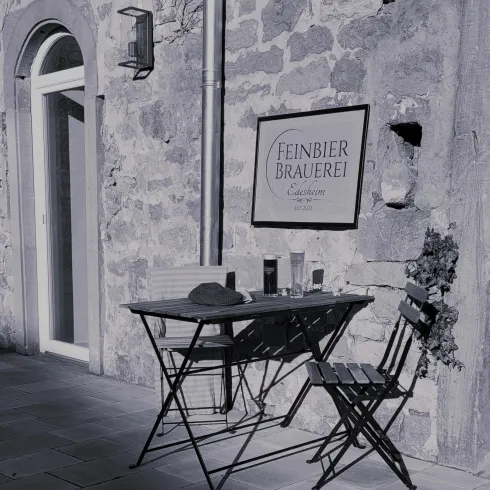 Ein kleiner Tisch mit zwei Stühlen steht vor einer steinernen Wand. An der Wand hängt ein Schild mit der Aufschrift „Einber Schönheit“.