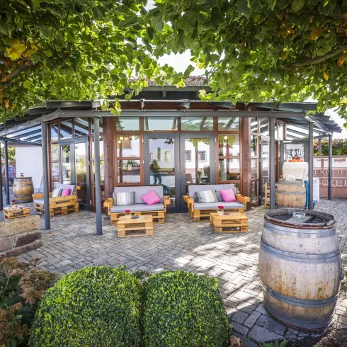 Ein einladendes Gebäude mit einer modernen Glasfront und bequemen Sitzgelegenheiten im Freien. Umgeben von schönen Pflanzen und Fässern, strahlt der Ort eine gemütliche Atmosphäre aus.