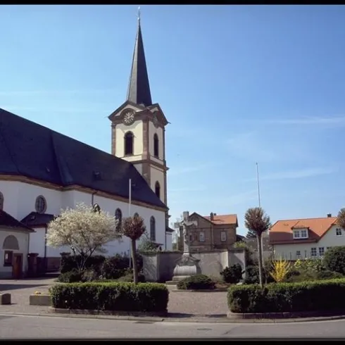 Eine Kirche mit einem hohen Turm steht im Vordergrund. Im Hintergrund sind Häuser und ein klarer blauer Himmel zu sehen.