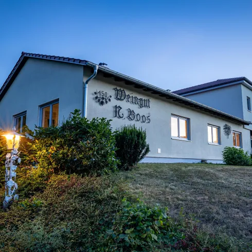 Ein modernes, helles Gebäude mit einem gepflegten Garten und einer warmen Außenbeleuchtung. Im Vordergrund ist ein Schild mit dem Namen "Wangyi E. Bodo" zu sehen.