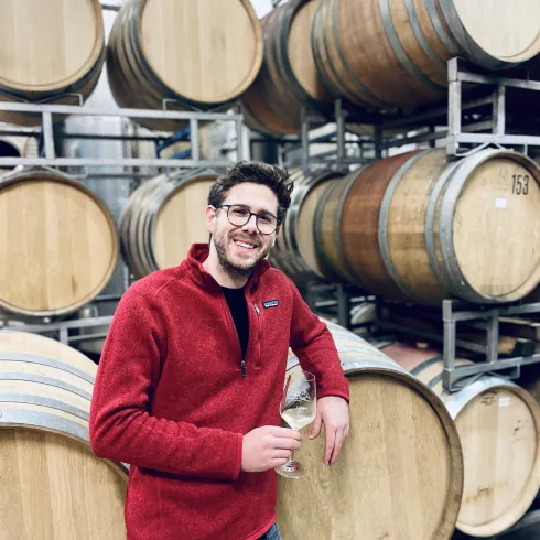 Ein junger Mann steht lächelnd vor Weinfässern in einem Weinkeller. Er hält ein Glas Wein in der Hand und trägt einen roten Pullover.