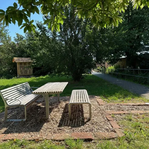 Ein ruhiger Garten mit einem Tisch und Bänken aus Holz. Umgeben von Bäumen und einer Spazierweg im Hintergrund.