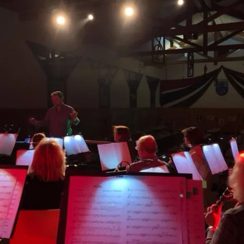Ein Musiker leitet ein Orchester in einer gut beleuchteten Halle. Die Musiker sitzen mit Notenblättern vor sich und konzentrieren sich auf die Aufführung.