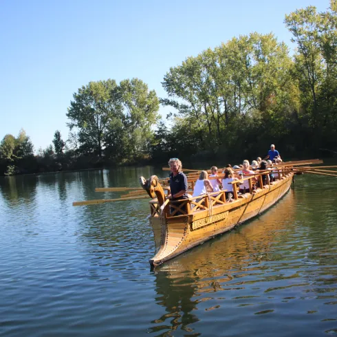 Eine Gruppe von Menschen fährt in einem Holzboot auf einem ruhigen Gewässer. Umgeben von grünen Bäumen und klarem Himmel genießen sie die Natur.