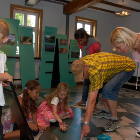 Eine Gruppe von Kindern und Erwachsenen in einem Raum. Sie beschäftigen sich miteinander und erkunden verschiedene Materialien auf dem Boden.