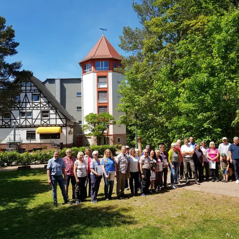 Eine Gruppe von Menschen steht im Freien vor einem Gebäude, umgeben von grünen Bäumen. Es ist ein sonniger Tag und die Atmosphäre wirkt freundlich und einladend.