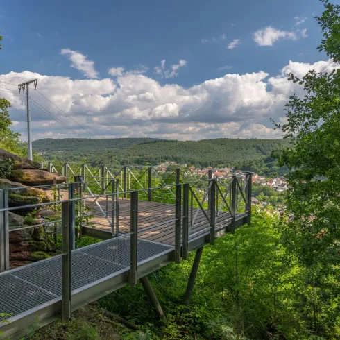 Eine Aussichtsplattform umgeben von Bäumen bietet einen Blick auf die Landschaft. Der Himmel ist blau mit einigen Wolken.
