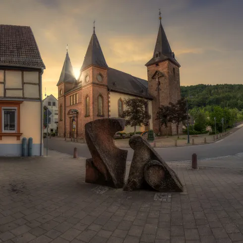 Eine alte Kirche mit zwei Türmen steht im Vordergrund, umgeben von modernen und historischen Gebäuden. Im Vordergrund befindet sich eine künstlerische Skulptur auf dem gepflasterten Platz.