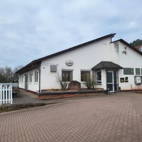 Ein modernes Gebäude mit einem Eingang und Fenstern. Der Parkplatz vor dem Gebäude ist gepflastert und es gibt einige Pflanzen in der Umgebung.