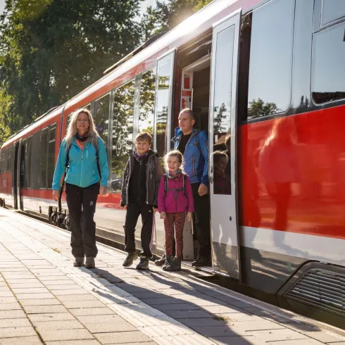 Eine Familie steigt an einem Bahnhof aus einem roten Zug aus. Die Sonne scheint und es ist eine grüne Umgebung zu sehen.
