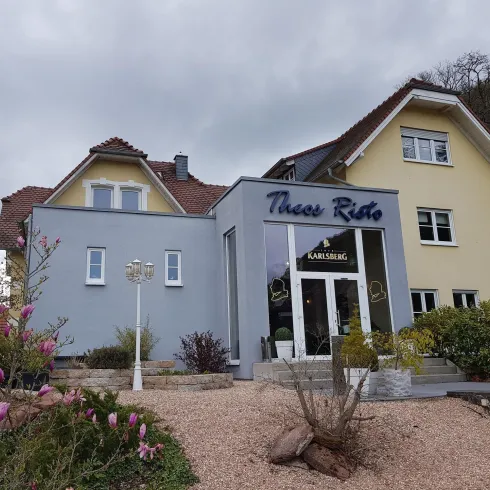 Eine moderne Villa mit zwei farblich unterschiedlichen Seiten, es ist das Restaurant "Theos Risto". Der Eingangsbereich ist einladend mit einem gepflegten Garten.