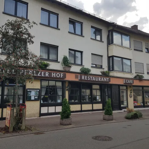 Ein Gebäude mit mehreren Geschäften im Erdgeschoss, darunter ein Restaurant und ein Café. Die Fassade ist in sanften Farben gehalten und es sind einige Pflanzen vor dem Gebäude zu sehen.