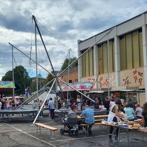 Ein lebhafter Marktplatz mit vielen Menschen und Ständen. Im Hintergrund stehen Fahrgeschäfte und ein großes Gebäude.