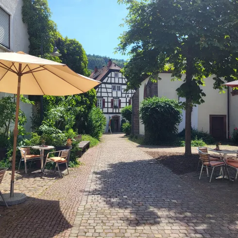 Eine idyllische Innenhof mit Bäumen und Sonnenschirmen. Es gibt gemütliche Sitzgelegenheiten und historische Gebäude im Hintergrund.