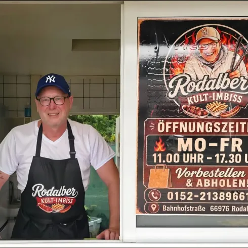 Ein freundlich lächelnder Mann steht am Fenster eines Imbisses. Im Hintergrund ist ein Schild mit den Öffnungszeiten und Informationen zu sehen.