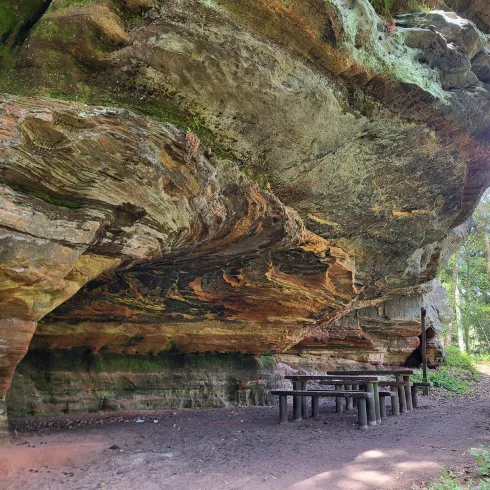 Eine beeindruckende Felsformation mit Überhang. Darunter befindet sich ein Tisch aus Holz und umgeben von Bäumen.
