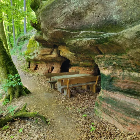Eine natürliche Felsformation mit einem kleinen Tisch und Bänken in einem Wald. Der Weg führt sanft an der Felswand entlang, umgeben von Bäumen und Laub.