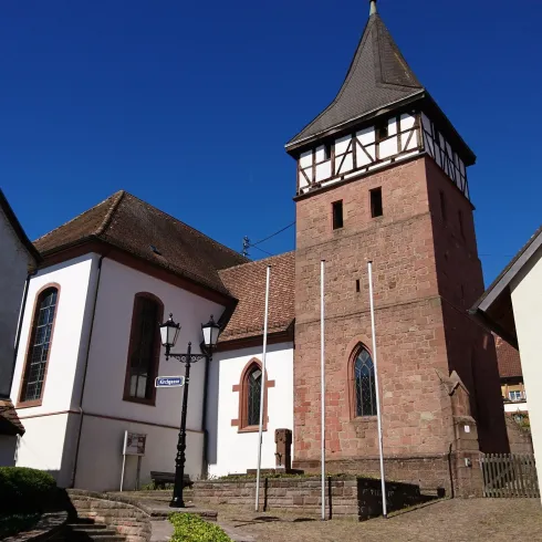 Ein historischer Turm mit einem spitzen Dach steht neben modernen Häusern. Der Himmel ist klar und blau.