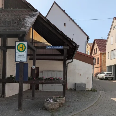 Eine Haltestelle an einer ruhigen Straße mit einem überdachten Wartestand. Im Hintergrund sind moderne Häuser sichtbar.