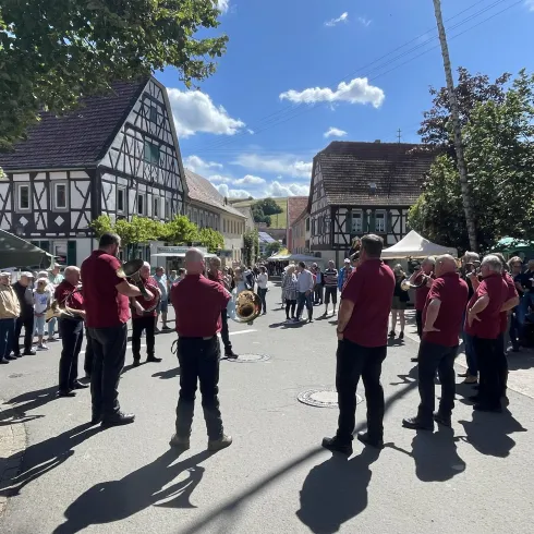 Eine Gruppe von Männern in roten Hemden steht auf der Straße, während sie einer Darbietung zusehen. Im Hintergrund gibt es ein traditionelles Fachwerkhaus und Bäume.