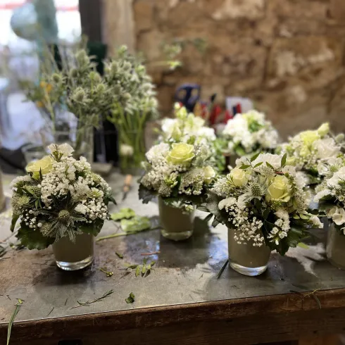 Auf dem Foto sind sieben kleine Blumensträuße in kleienen Töpfen.Das Ganze steht auf einem Tisch. Im Hintergrund stehen noch weitere Blummensträuße.