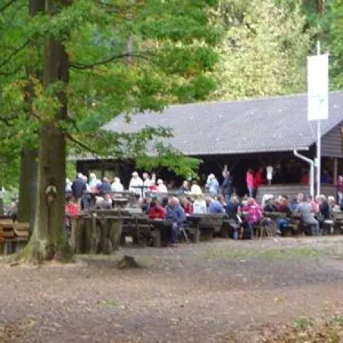 Ein gemütlicher Außenbereich mit langen Tischen und Bänken, besetzt mit Menschen. Im Hintergrund das schöne, holzige Gasthaus im Wald.