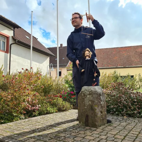 Ein Mann steht auf einem Stein und hält eine Marionette in der Hand. Im Hintergrund sind Blumen und Gebäude sichtbar.