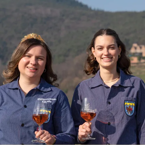 Zwei Frauen halten Gläser mit Wein und lächeln in die Kamera. Im Hintergrund sind sanfte Hügel und ein klarer Himmel zu sehen.