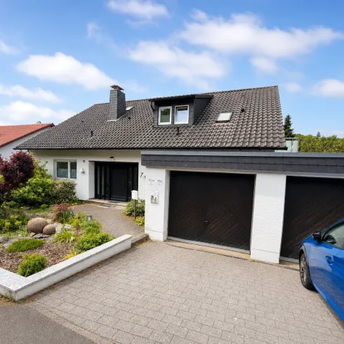 Ein modernes Haus mit einem begrünten Vorgarten und zwei Garagen. Ein blauer Wagen steht vor dem Eingang.