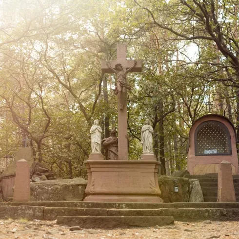 Ein ruhiger Ort im Wald mit einem großen Kreuz und zwei Statuen. Im Hintergrund ist eine kleine Kapelle zu sehen.