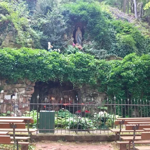 Eine kleine Kapelle inmitten von grünem Bewuchs in einem Wald. Vor der Kapelle stehen Bänke für Besucher.