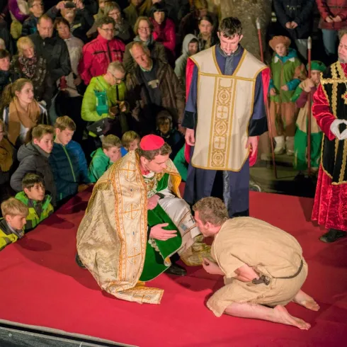 Eine lebhafte Aufführung mit Schauspielern in historischen Kostümen auf einer roten Bühne. Im Vordergrund kniet ein Darsteller vor einer weiteren Figur, während das Publikum zuschaut.