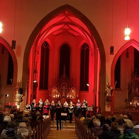 Eine Gruppe von Sängern tritt in einer festlich beleuchteten Kirche auf. Die Zuschauer sitzen in einem vollen Gottesdienstsaal unter roten Lichtern.