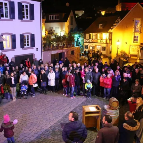 Eine große Menschenmenge versammelt sich auf einem Platz bei Nacht. Umgeben von festlich beleuchteten Häusern, scheinen die Menschen eine Veranstaltung zu genießen.