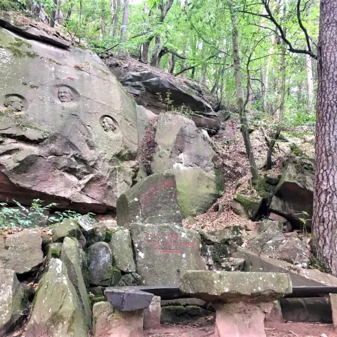 Eine mystische Felsformation im Wald mit eingravierten Symbolen. Darunter befindet sich eine Bank aus Stein, umgeben von Bäumen und lichter Vegetation.