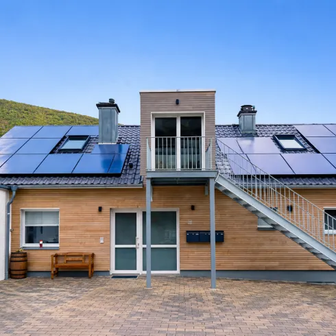 Ein modernes Haus mit Solarpanelen auf dem Dach und einer Außentreppe. Die Fassade ist teilweise in Holzoptik gestaltet.