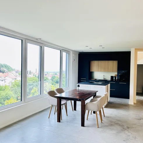 Ein modernes Wohn- und Esszimmer mit großen Fenstern und Blick auf die Landschaft. Der Tisch ist aus Holz, umgeben von gepolsterten Stühlen.