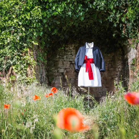 Ein traditionelles Kleid mit einer blauen Jacke und einem roten Gürtel steht in einer grünen Umgebung. Im Vordergrund blühen rote Mohnblumen.